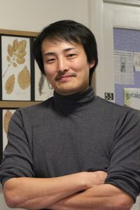 Kiwamu Tanaka stands with his arms crossed in front of a photo of leaves.
