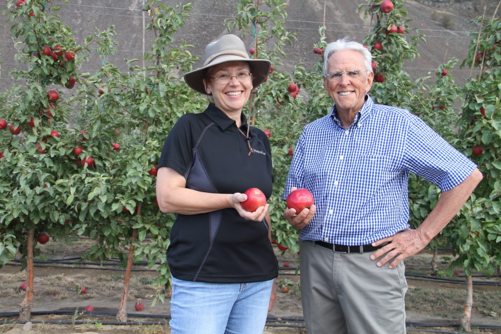 Evans. left, and Barritt hold red apples in front of a wall of trees.