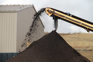Black compost tumbles off an inclined yellow conveyor into a dark pile. A shed and hill stand in the background.