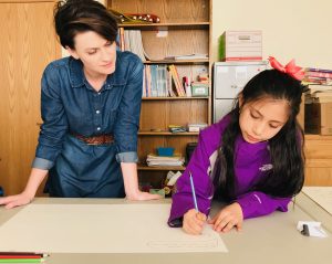 A standing teacher at left overlooks a student creating artwork at a table.