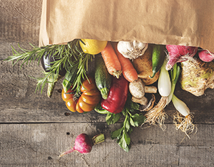 Fresh vegetables spill from a shopping bag, sitting on its side, onto a wooden counter.