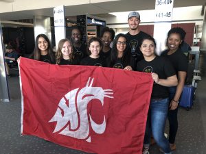 Nine students wearing identical black shirts hold a WSU flag in an airport.