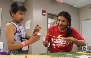 Boy, left, and adult helper, right, unfurl a ribbon above a craft table.