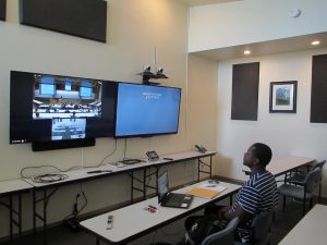 A student sits at a desk looking at 2 big monitors on the wall. 1 monitor shows a classroom in another city.