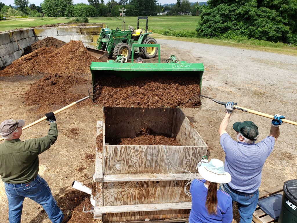2 people use pitchforks to get small woodchips out of a tractor's front loader and into a wood crate. 