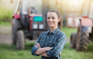 Woman farmer standing on farmland with crossed arms and tractors in background