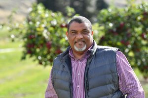 Dean Wright stands in an apple orchard looking at the camera. A tree laden with red apples is out of focus in the background.