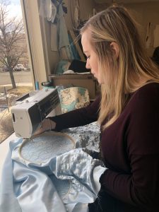 Blonde woman using a sewing machine by a window. She is sewing a blue dress.