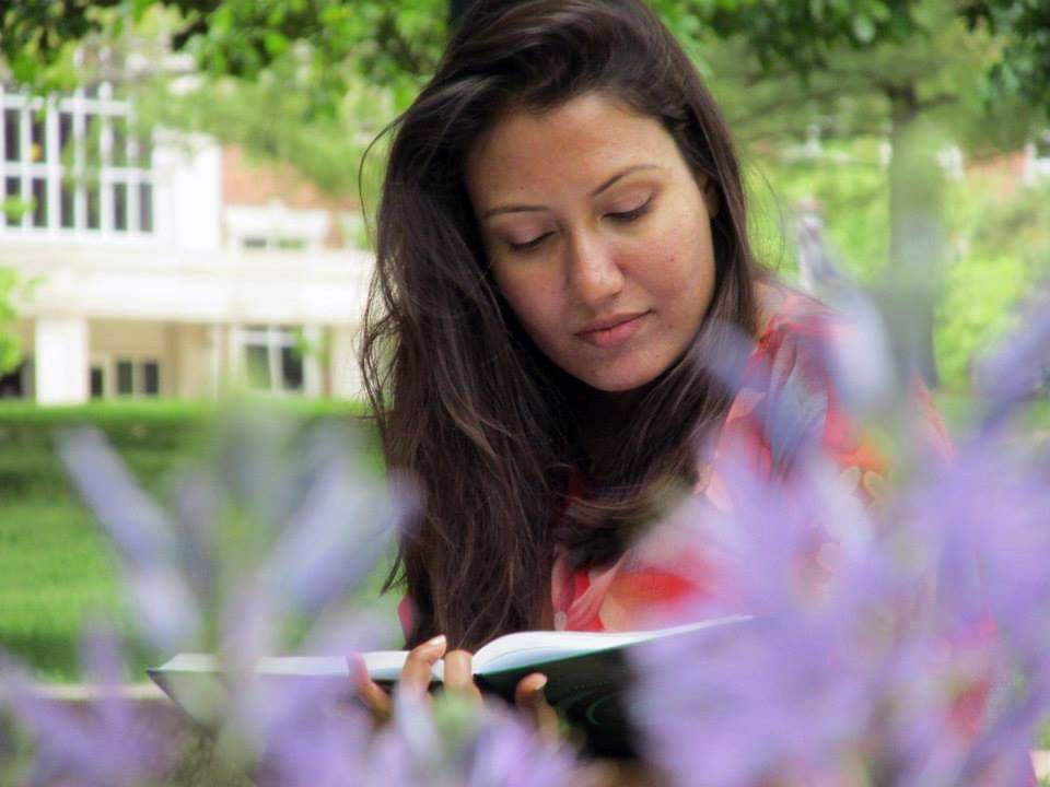 A young woman reading a book next to flowers on a campus lawn.