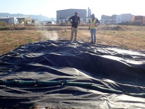 A black tarp on a field with steam seeping out from the edges. Two people stand nearby observing.