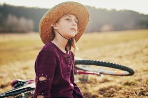 A young girl with a sun hat on and a maroon shirt gazes at the sun against the backdrop of a forest with a golden field in front of it.