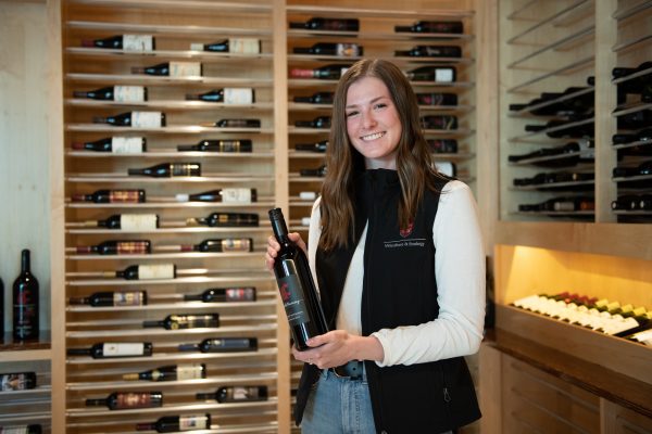 A young brunette woman in a black vest poses in front of a rack of wine with a bottle of wine.