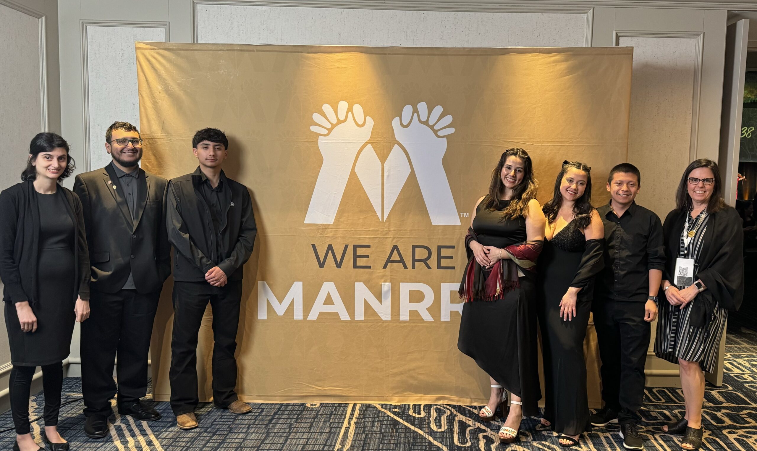 A group of seven people, three on one side and four on the other, stands next to a banner that reads "We are MANRRS" with a symbol of two sets of hands interlocking.