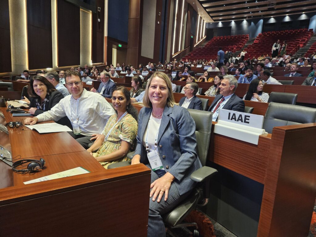 Four people sitting in chairs at a table look at the camera. Behind them are more seats with people sitting in them.
