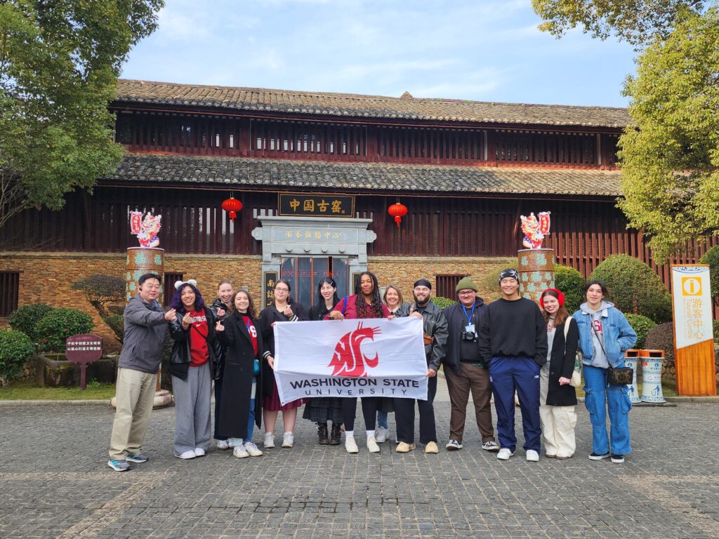 Group photo of 13 people holding a WSU flag outside of a traditional Chinese building.