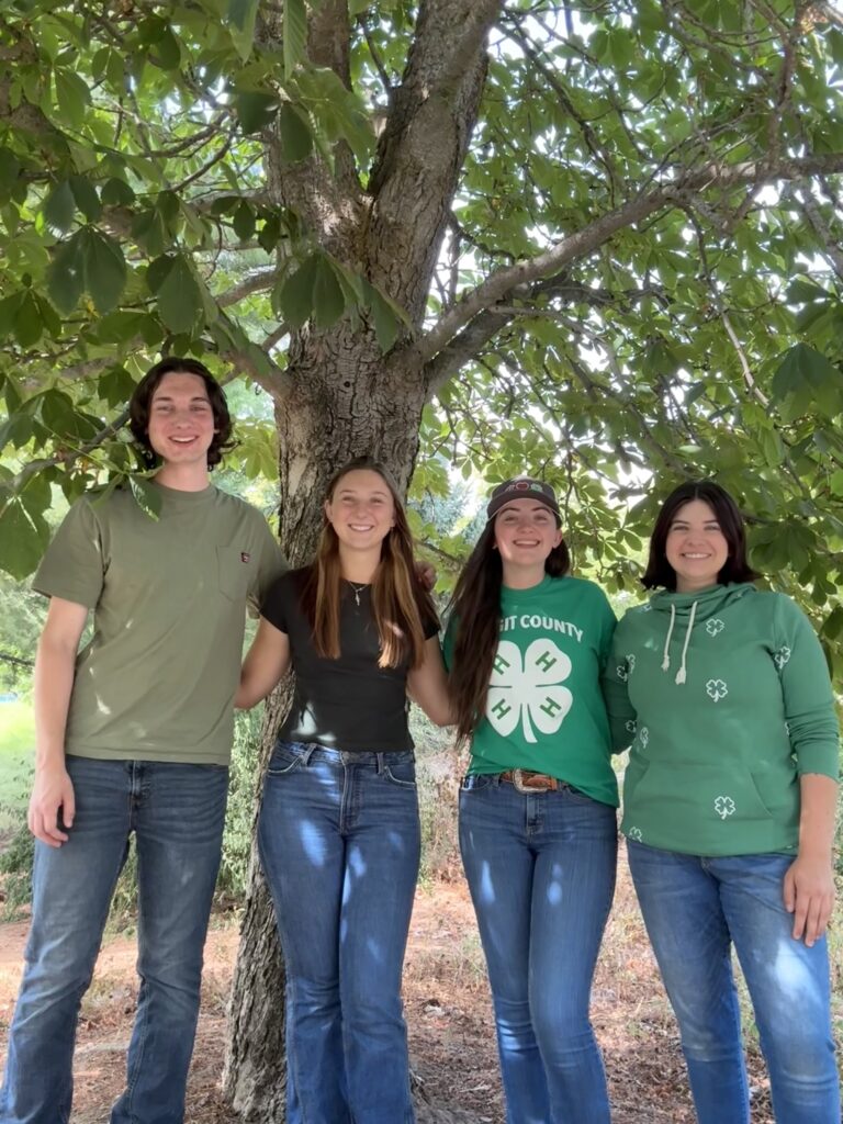 4 people stand together in front of a tree, with 2 of them wearing 4-H clothing.