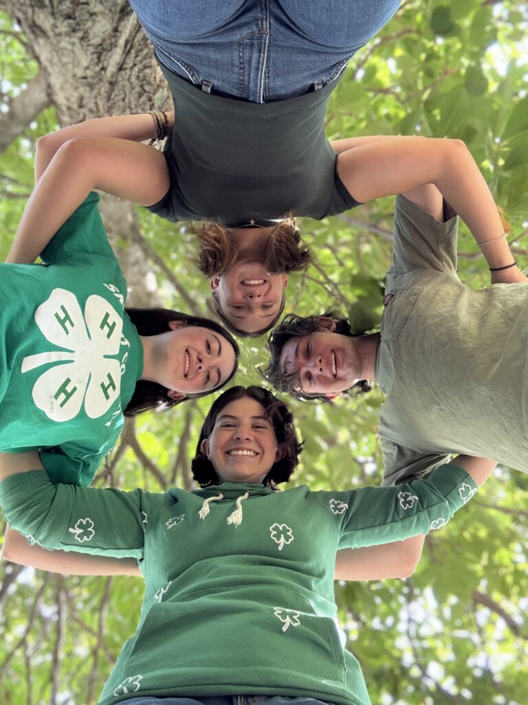 4 people stand in a circle looking down at the camera. They all have their hands around each other’s shoulders.