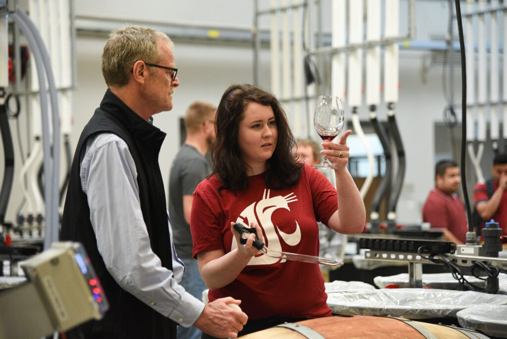Two people stand in the WSU Wine Science Center next to a wine barrel. One person holds up a glass with a small amount of red wine in it. In her other hand, she holds a glass pipette. Several other people stand in the background.
