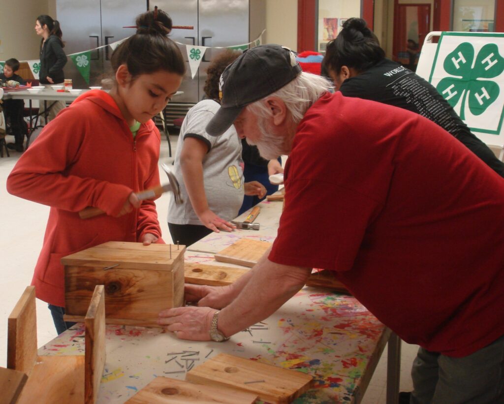 A youth hammers boards down while an adult holds the woodworking project at the base to help stabilize it.