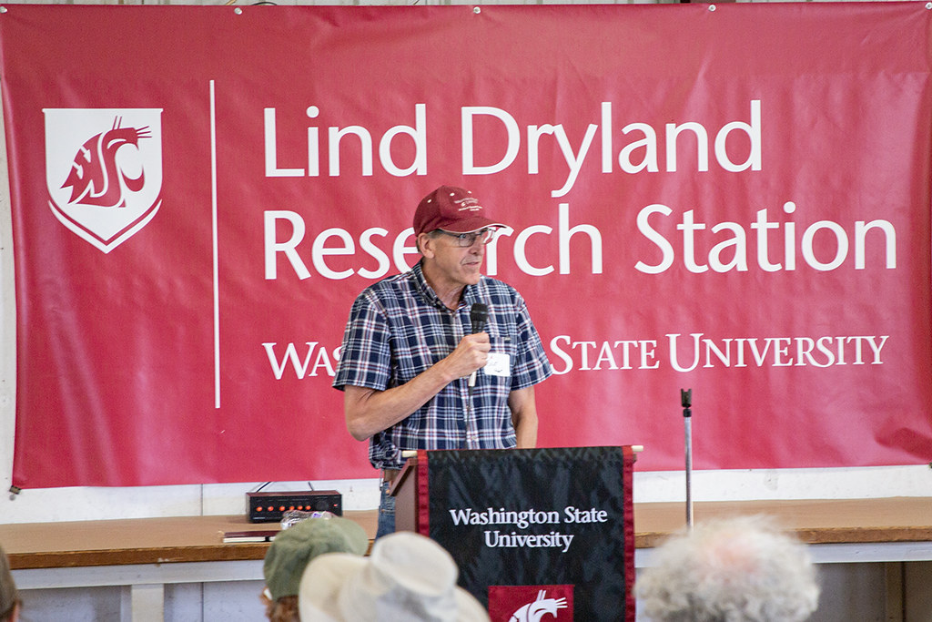 A person speaks into a microphone while standing at a podium. The backs of the heads of people in an audience are pictured. Behind the speaker is a sign with the WSU cougar head logo and the words "Lind Dryland Research Station Washington State University."
