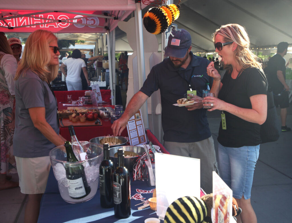 A person stands behind a table with bottles of wine and bowls with cheese samples on it. In front of the table are two people. One reaches into a bowl for a cheese sample, while the other places her cheese sample into her mouth.