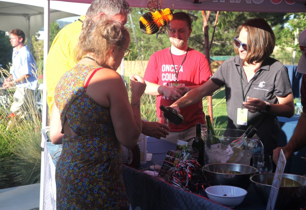 Two people stand behind a table, and two people stand in front of it. One of the people standing behind the table pours wine into the glass of a person standing in front of the table. On the table are metal bowls of cheese and meat samples, bottles of wines, pom poms, a jar of honey and other WSU-related items.