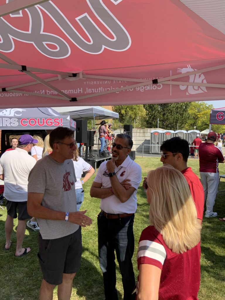 People in WSU Cougar-themed attire talk together under a tent at a sunny campus tailgate event.