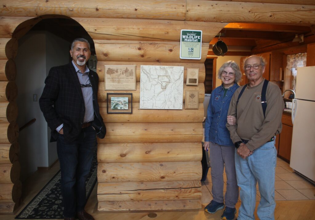 Three people (one on the left and two on the right) stand inside of a log cabin on either side of signs on the wall. One is a topographical map of the property, one reads "Casa Becca del Norte Tree Farm" with an illustration of a cabin near trees. Another sign reads "Certified Wildlife Habitat." There is also a small painting of a forested property, and two other award plaques.