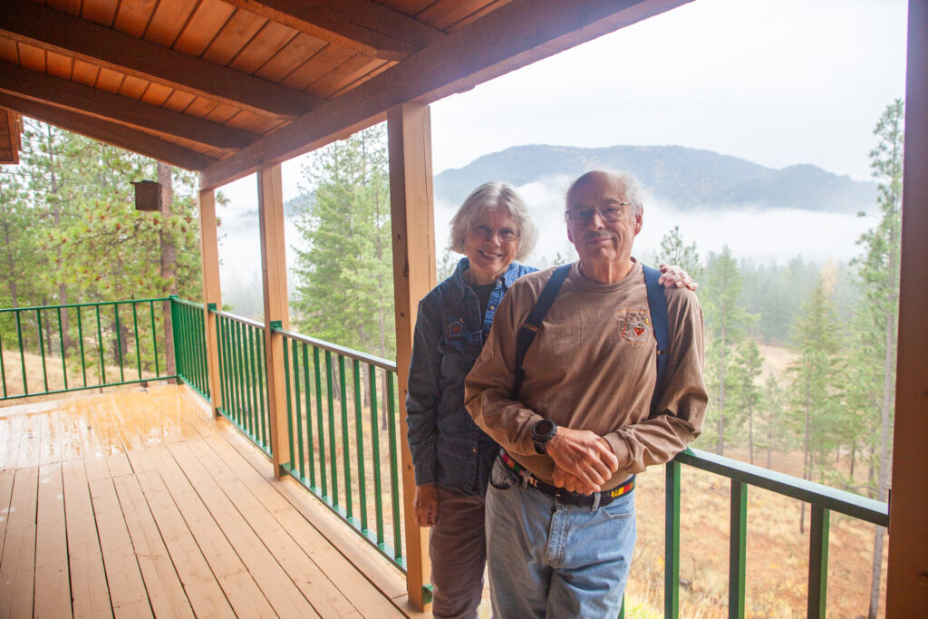 Two people stand on the covered deck of a log cabin. Behind them are trees and forested hills partially covered in clouds.