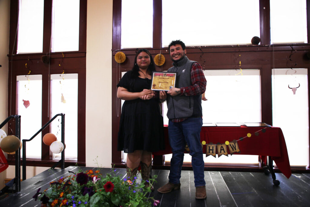 Two people stand together on a stage holding a framed award.