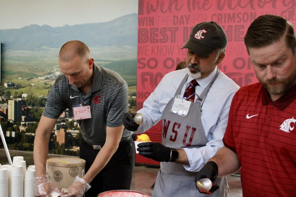 Dean Khosla, at center between two volunteers, scoops ice cream at a table; behind is a red banner and scenic photo of farmland and hills. 