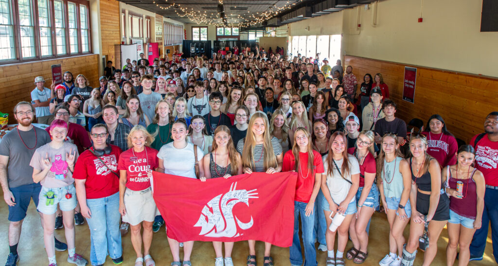 A large group of students, staff, and faculty inside Ensminger Pavilion. Several people in the front of the group hold a red flag with a white WSU cougar head logo on it.