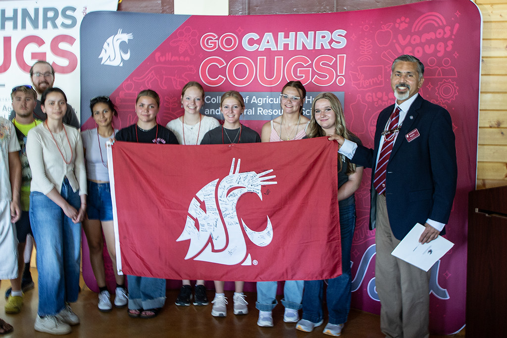 Dean Khosla, right, holds the crimson WSU banner with a group of students. A background display reads Go CAHNRS Cougs.