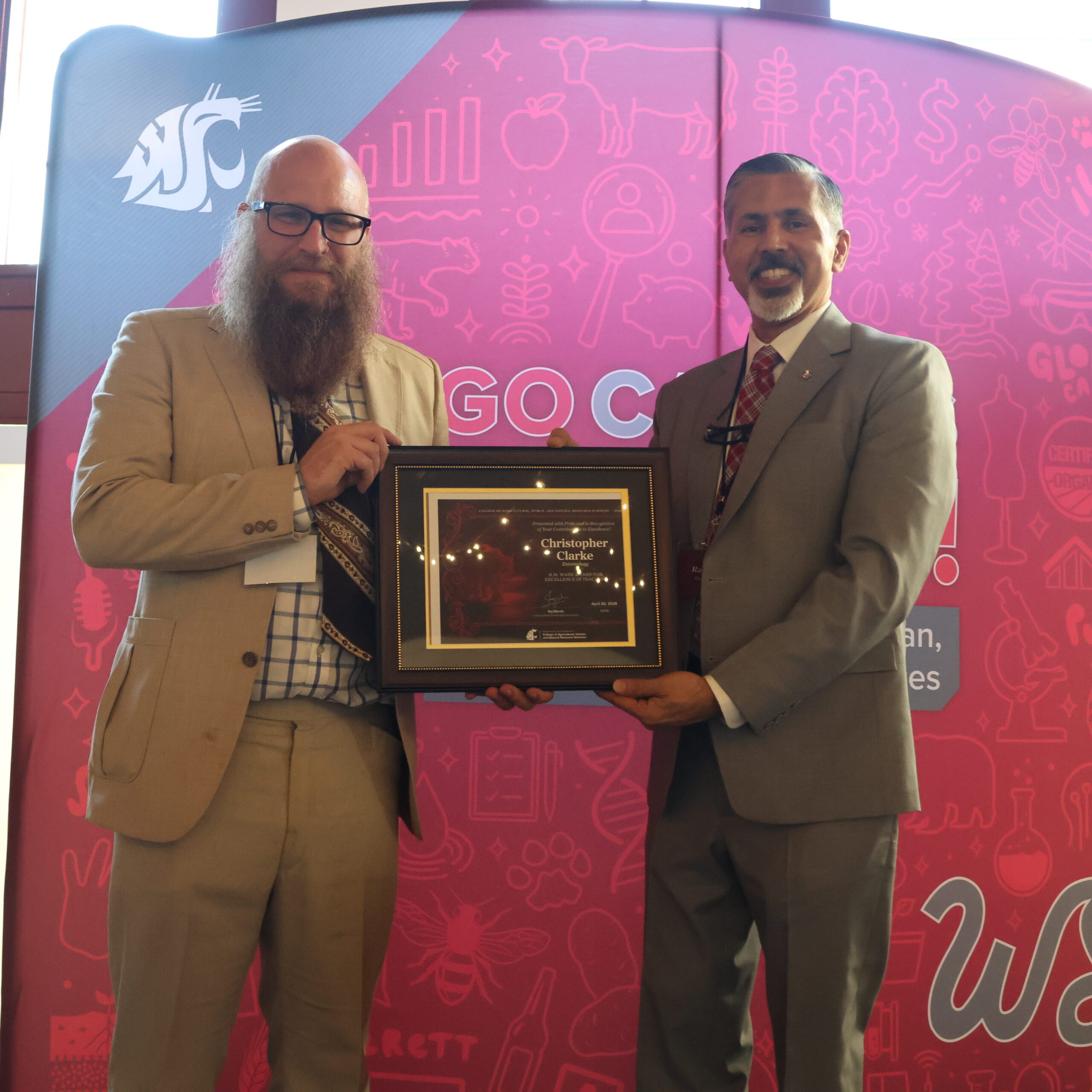 Award recipient Christopher Clarke, left, holds award certificate on stage in front of WSU-themed backdrop. CAHNRS Dean Raj Khosla stands at left, helping hold the certificate.