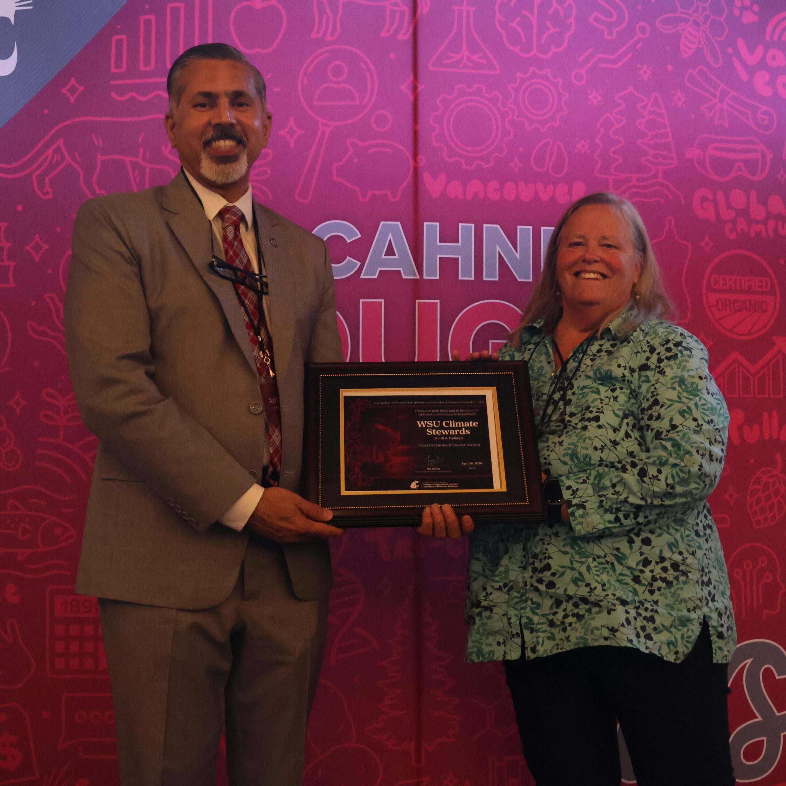Karen Lewis, rights, holds an award certificate (for the WSU Climate Stewards Team) on stage in front of WSU-themed backdrop. CAHNRS Dean Raj Khosla stands at left, helping hold the certificate.