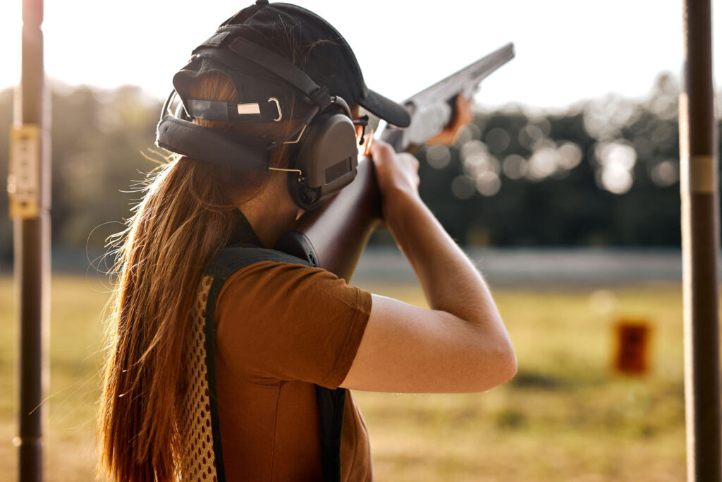 The back of a person with long hair wearing ear protection and a hat. The person points a gun out into a field with a blurred object.