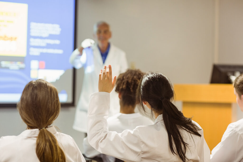 A student in a classroom raises her hand as a teacher at the front points at her. Everyone is wearing white lab coats.