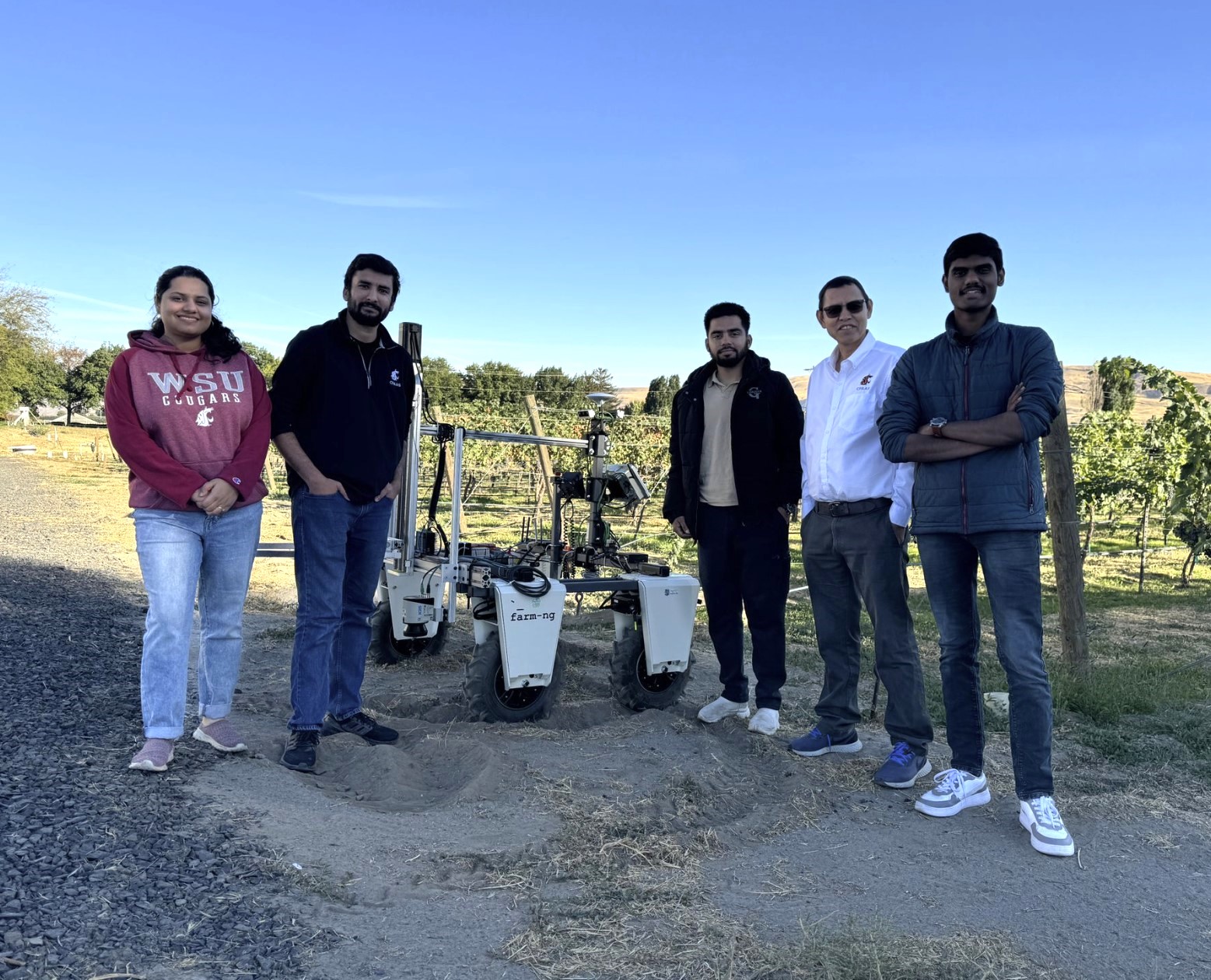 A group of five people stands next to a robot. Behind them is a vineyard.