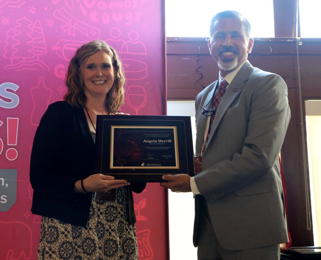 Award winner Angela Merrill, left, poses with a framed award certificate with Dean Raj Khosla, right, in front of windows and a crimson backdrop.