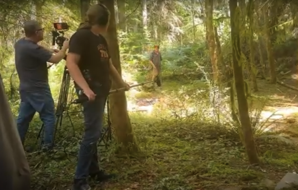 2 people stand in a woods, one working a camera while the other holds a boom microphone. A third person is in the background, walking toward the camera.