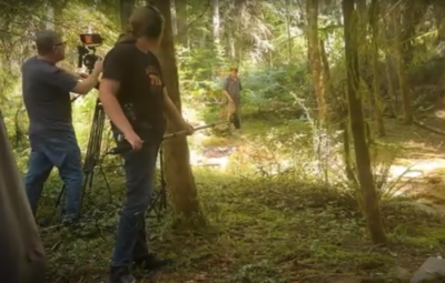2 people stand in a woods, one working a camera while the other holds a boom microphone. A third person is in the background, walking toward the camera.
