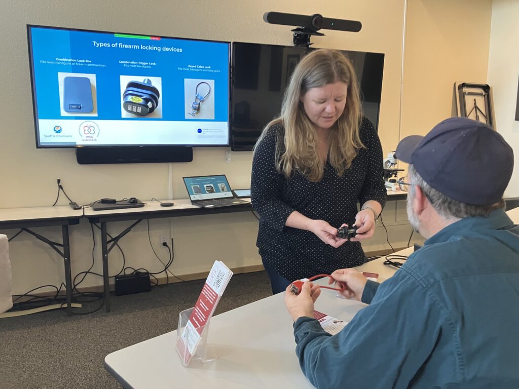 A person stands at a table, holding a firearm locking device. Another person is seated at the table, facing the person who is standing. Behind them is a TV screen displaying info about different types of firearm locking devices.