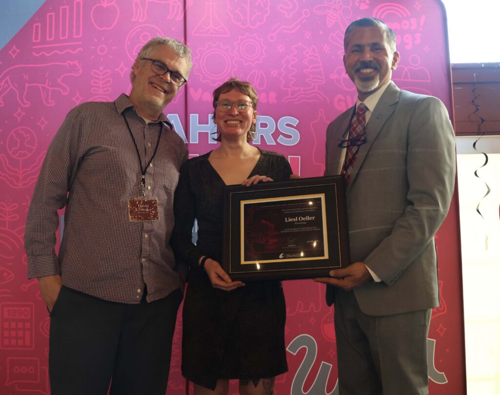 Award recipient photo- recipient Liesl Oller holds award at center, flanked by colleague David Crowder, left, and emcee Raj Khosla, right.