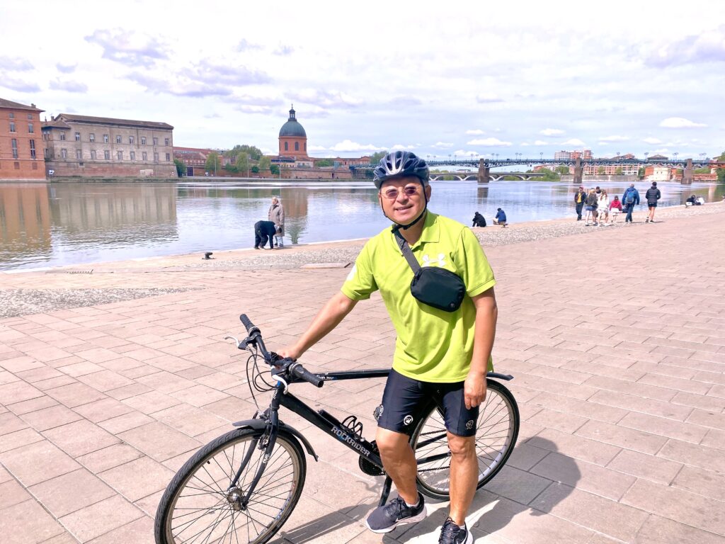 Yang, in cycling gear with bicycle, smiling in an urban riverside scene.