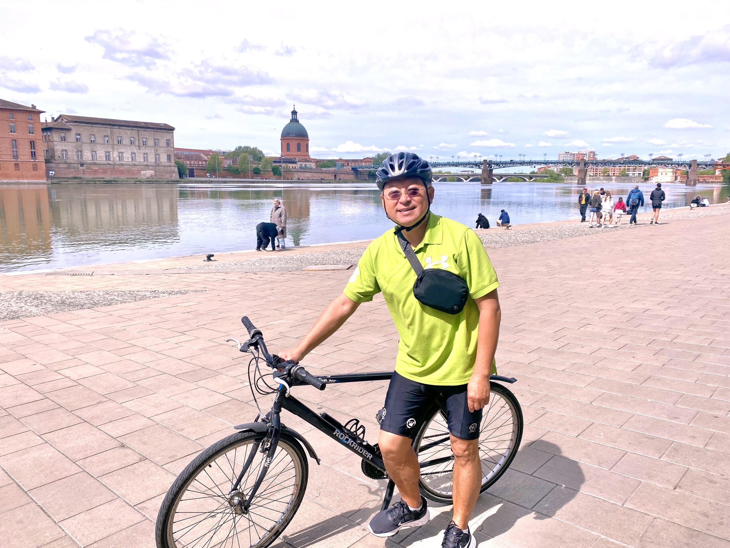 Yang, in cycling gear with bicycle, smiling in an urban riverside scene.
