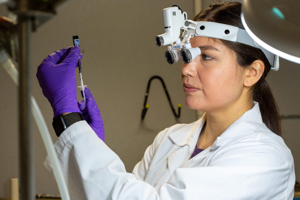 A person wears a lab coat and special glasses to look at a small vial of liquid as she withdraws material from it into a syringe.