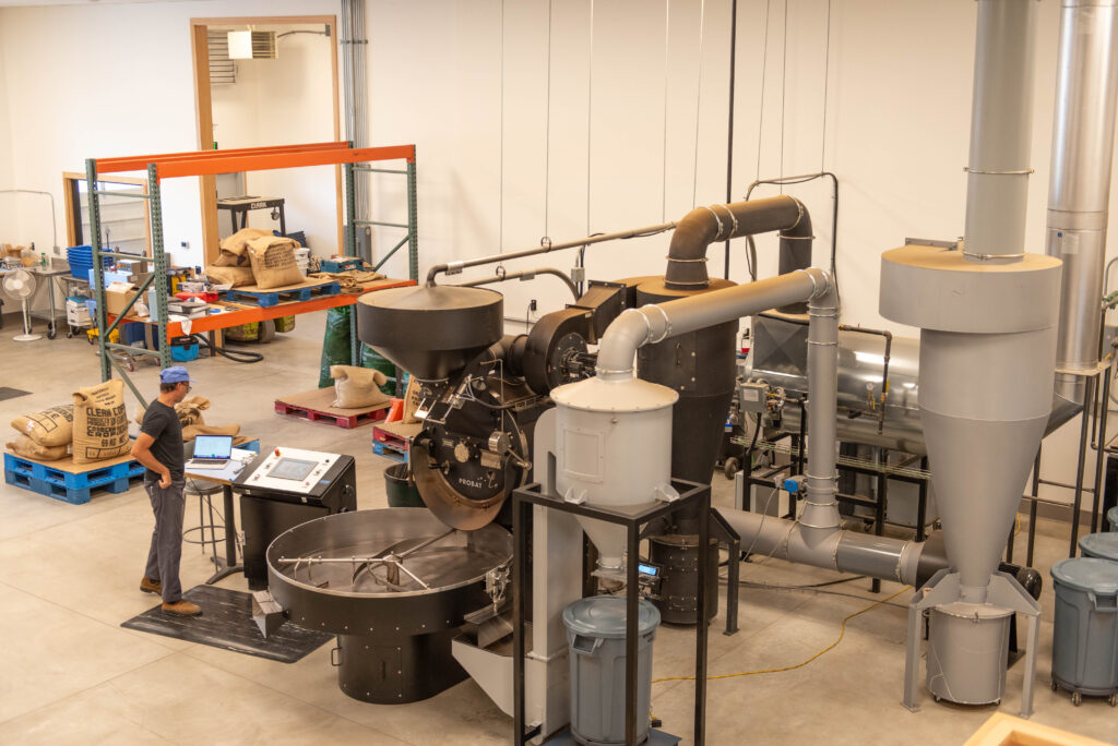 Industrial coffee roasting machinery in warehouse space. A person surveys the controls, and large sacks of coffee beans in burlap are on racks and pallets.