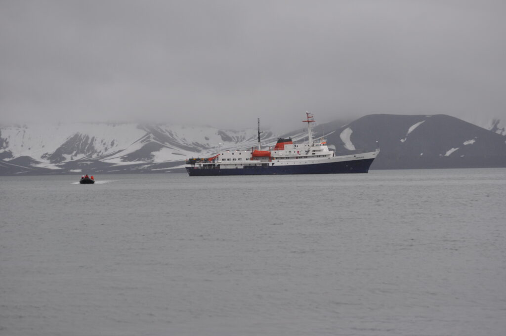 A large boat sits on calm water with large snow-covered mountains behind it.