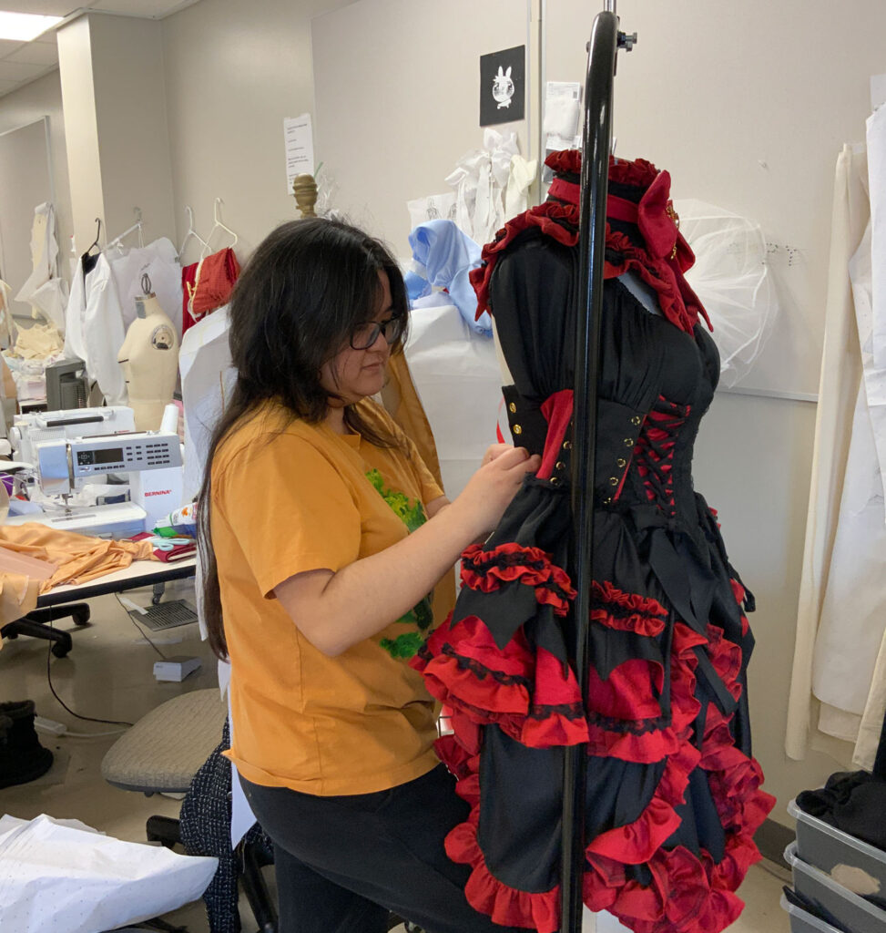 Britney Nguyen works on a black and red dress with lots of ruffles, layers, and a lace-up corset bodice. She's in a room with mannequins, sewing machines, and scraps of fabric.