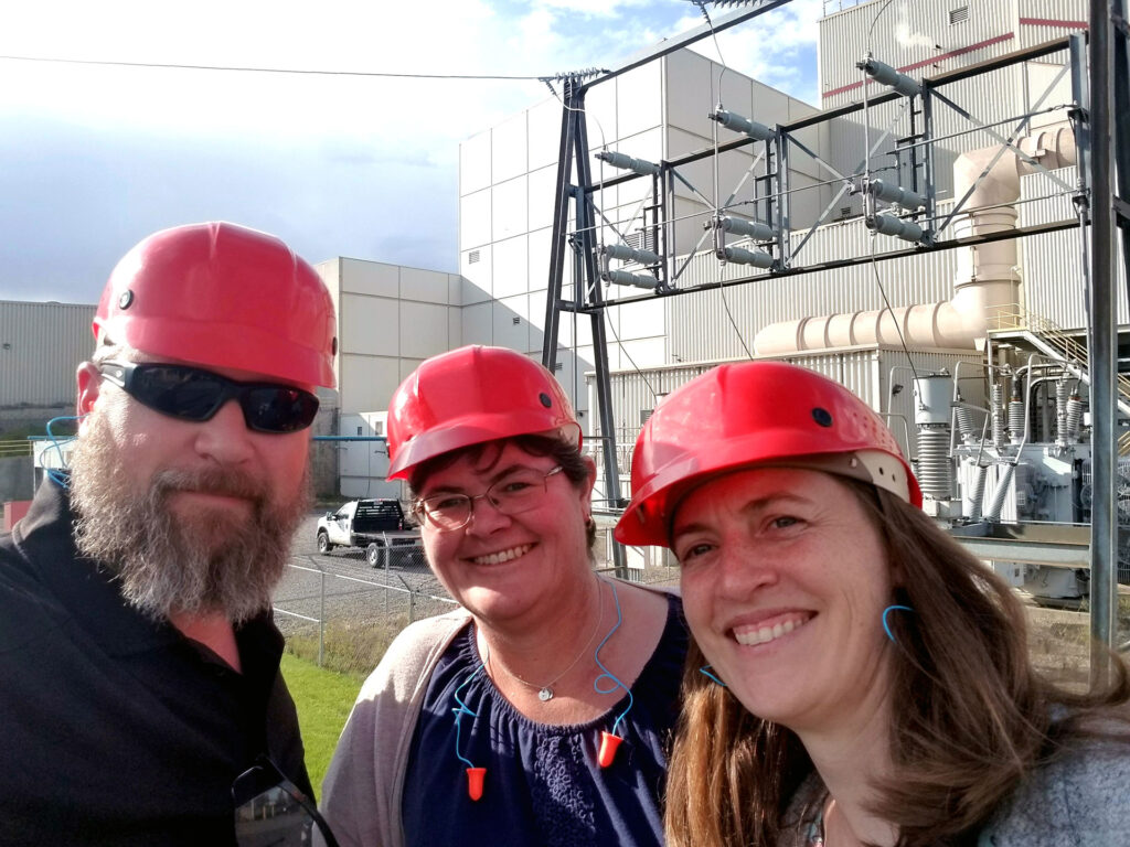 3 people wearing orange hard hats stand outside an industrial-looking facility.
